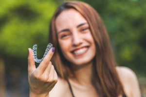 Woman smiling and holding up Invisalign tray with tree-esq background