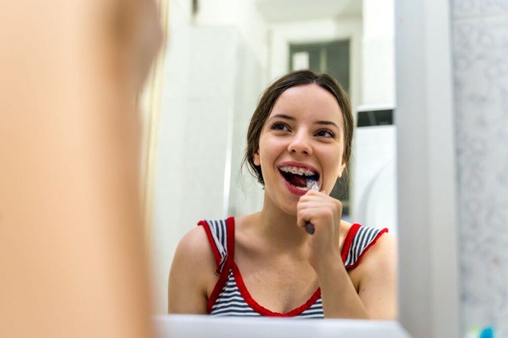 A teenage girl with braces brushing her teeth in a mirror.